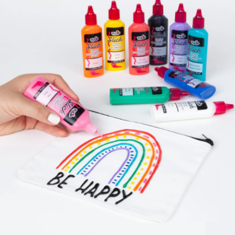 a hand hold a tube of puffy paint. There is a zipper pouch with a puffy paint rainbow an the words Be Happy under the rainbow. Puffy paint tubes are in the background. 