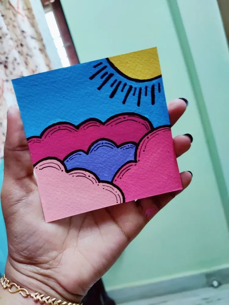 Hand holding a tiny painting of pink clouds, blue sky and a sun in the corner.